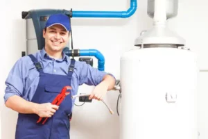 A person in blue overalls and a cap holds plumbing tools next to a water heater.