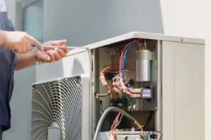 A technician repairs an outdoor air conditioning unit, using a screwdriver to adjust the wiring and components inside.