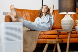 A woman relaxes on a brown leather couch with her feet up, next to a lamp and a dehumidifier. She is smiling with her eyes closed.