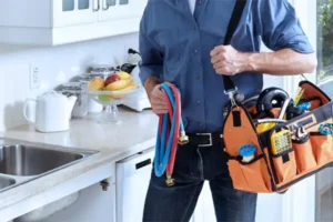 Person holding plumbing tools and an orange tool bag in a kitchen near a sink, with fruit and kitchen items in the background.