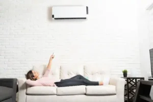 Person lying on a sofa in a white room, using a remote to control an air conditioner mounted on a brick wall.