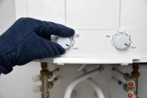 A gloved hand adjusts the settings on a water heater, turning the dial to high. Various pipes and connectors are visible below the device.